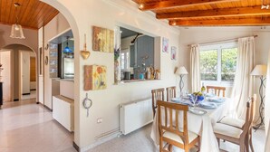 Ein Essbereich mit einem Holztisch, gedeckt für vier Personen, mit Stühlen an beiden Seiten. Im Hintergrund ist eine Küche erkennbar, durch ein Fenster ist Grün anzusehen.