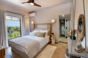 A bedroom with a large bed, a ceiling fan, an air conditioning unit, a bedside table with a vase, and a view of greenery outside. - Dimitris Cottage - Two Bedroom Villa, Sleeps 4 (Apergatika)