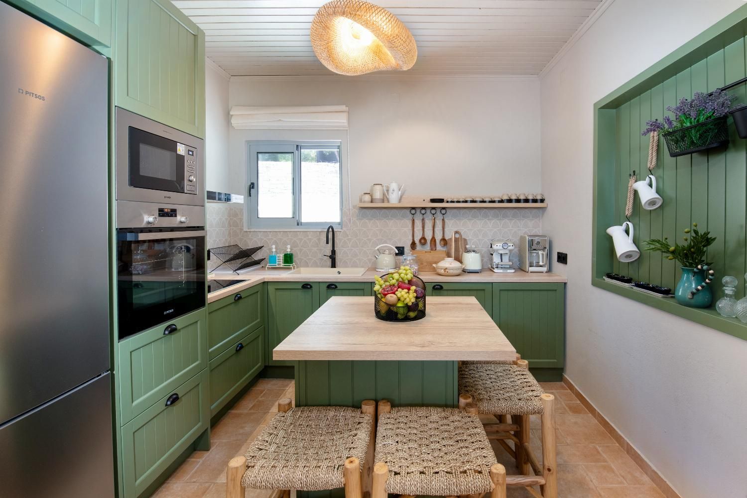 A kitchen with green cabinets, a stainless steel refrigerator, a wooden island with stools, and a hanging light fixture.