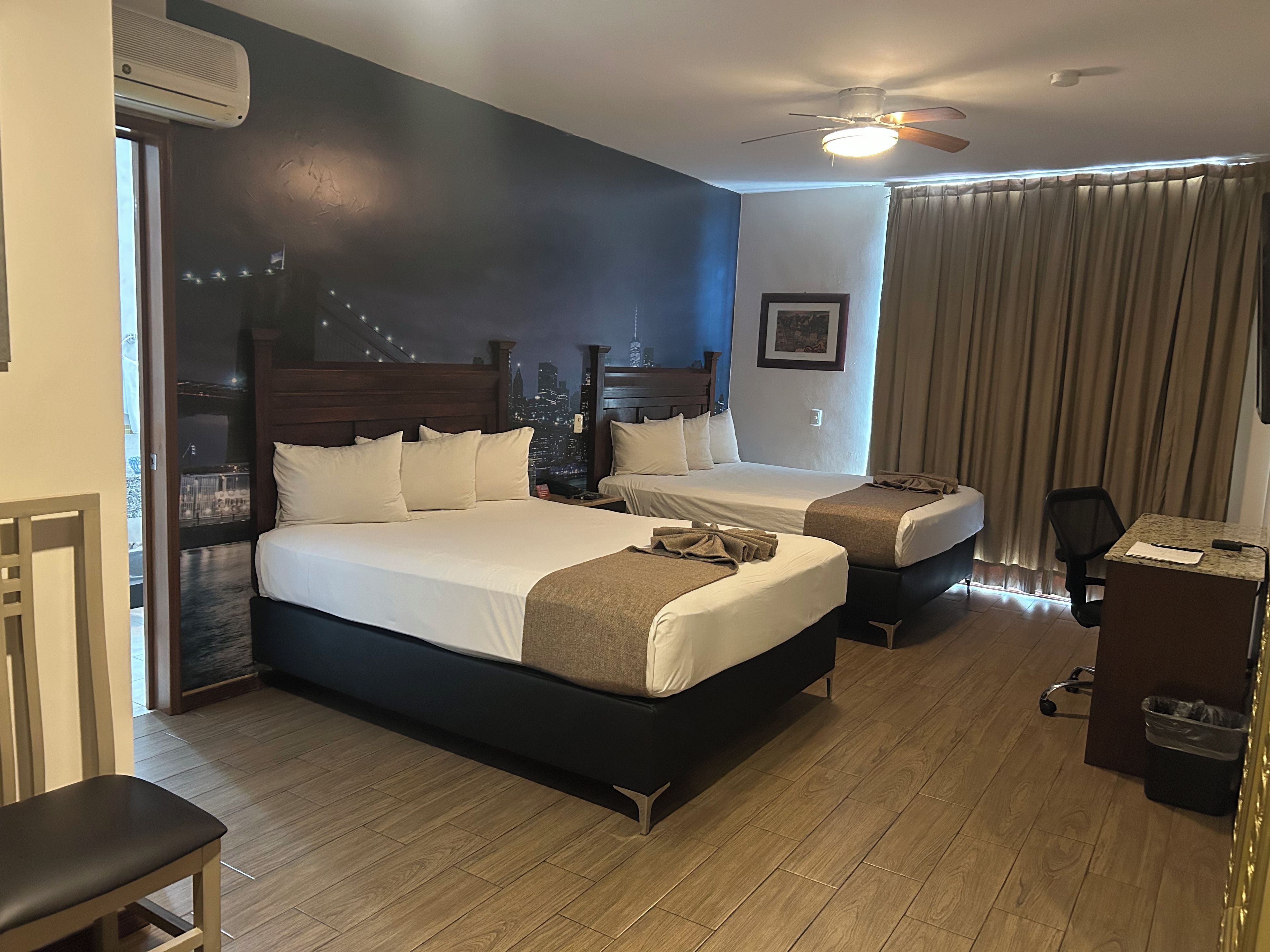 A hotel room with two beds, a desk, a chair, a ceiling fan, and a view of the cityscape.