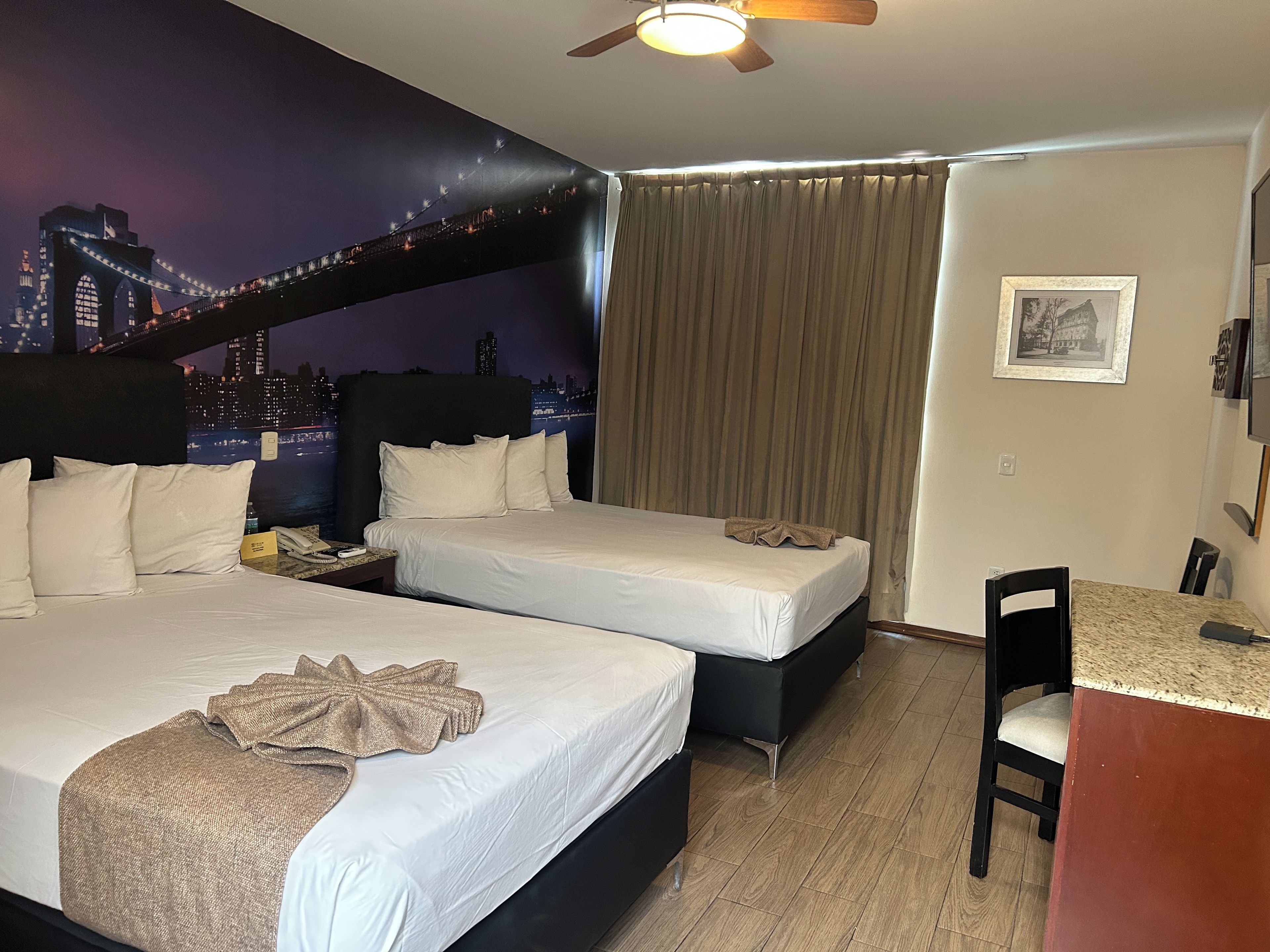 Ein Hotelzimmer mit zwei Betten, einem Schreibtisch, einem Stuhl, einem Deckenventilator und einem Wandgemälde mit Stadtansicht.