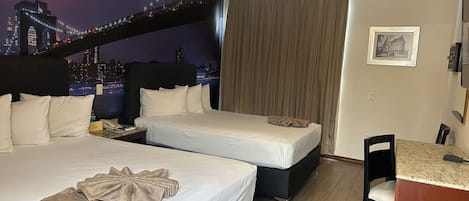 A hotel room with two beds, a desk, a chair, a ceiling fan, and a cityscape mural on the wall.
