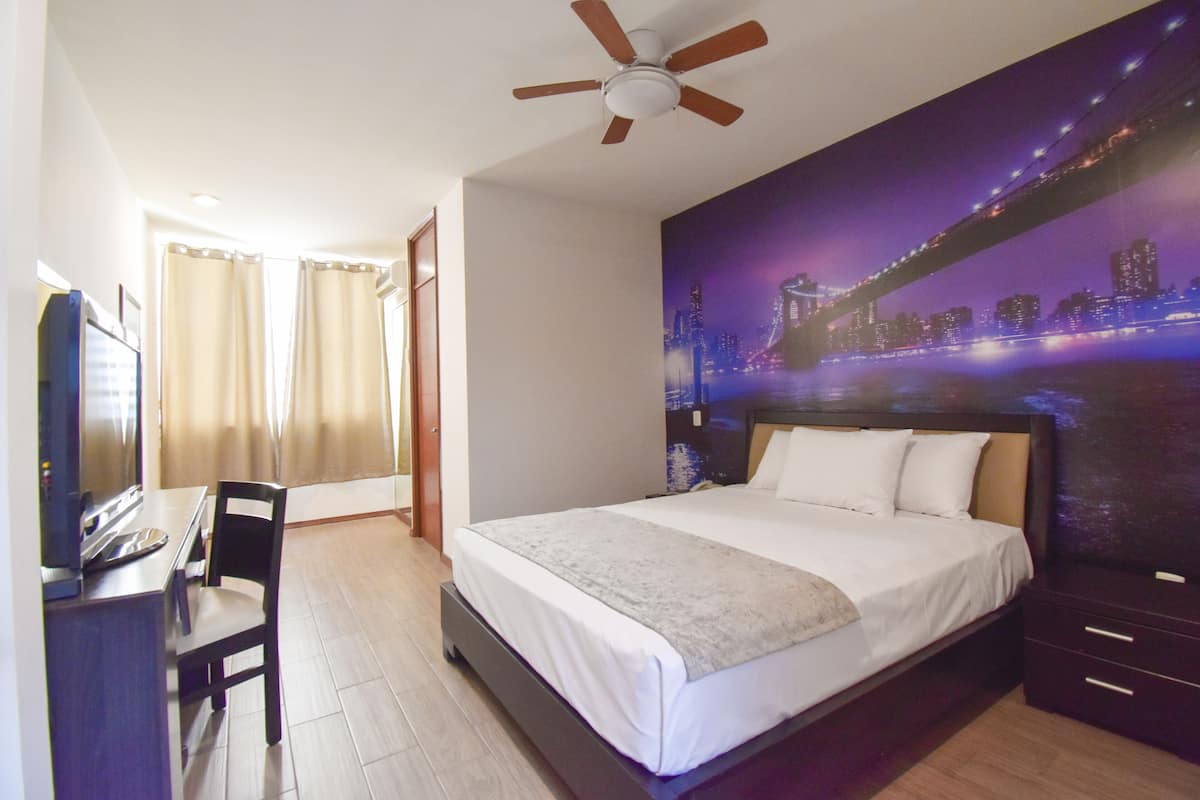 A hotel room with a bed, a desk, a chair, a ceiling fan, and a large cityscape mural on the wall.