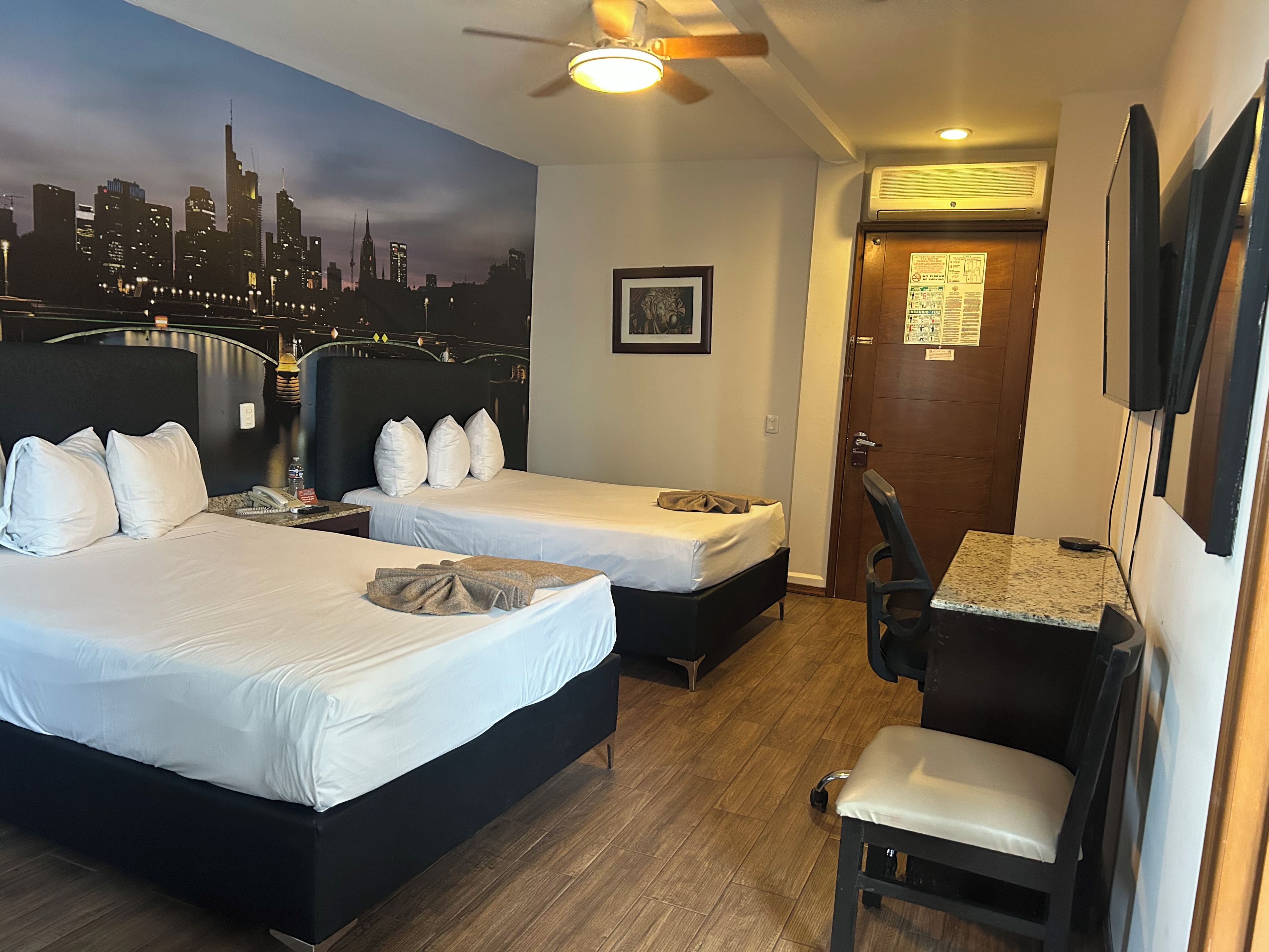 A hotel room with two beds, a desk, a chair, a cityscape mural, and a ceiling fan.