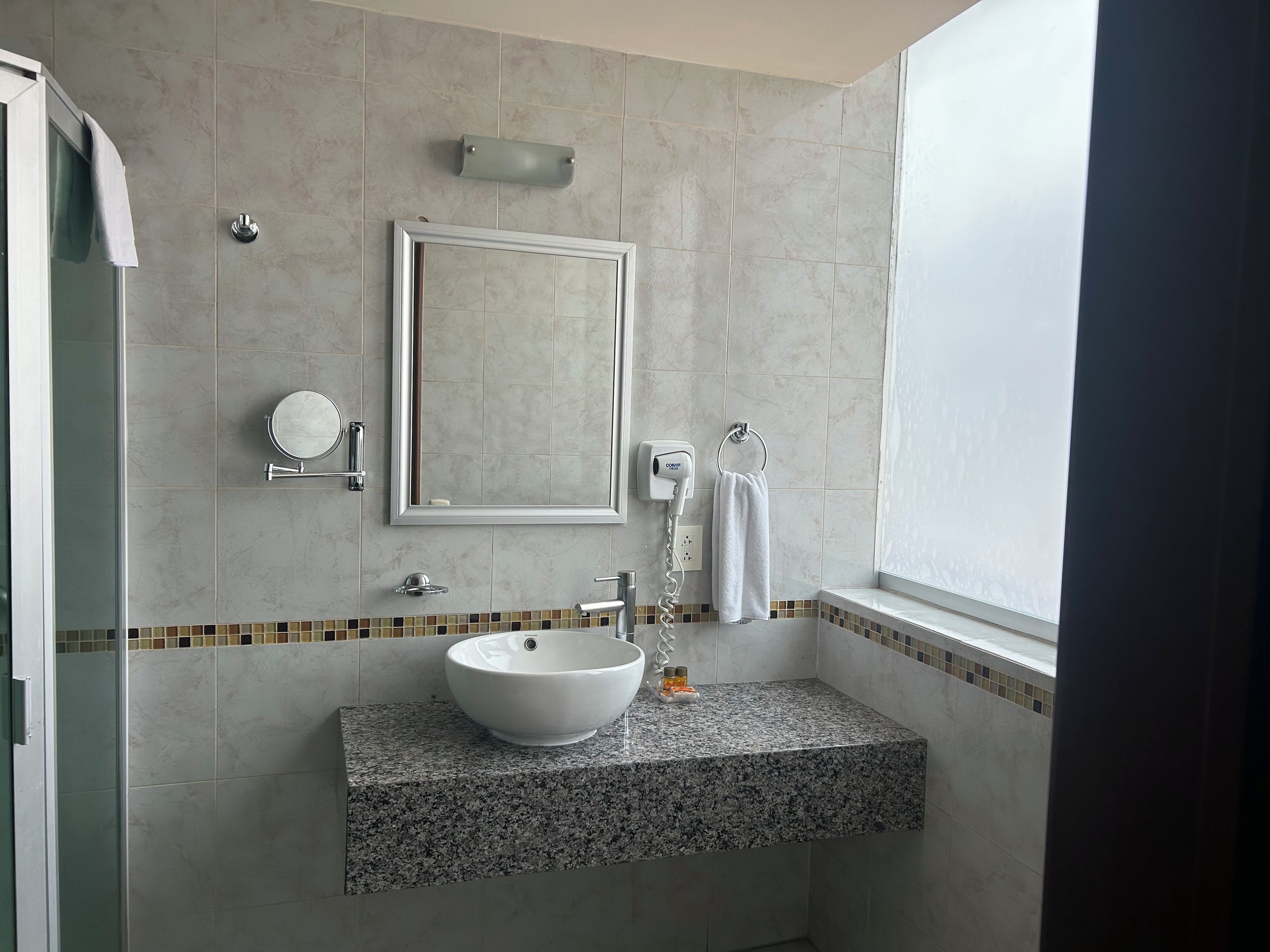 Une salle de bain moderne avec un comptoir en granit, un lavabo sur colonne, un miroir, un porte-serviettes et une fenêtre.