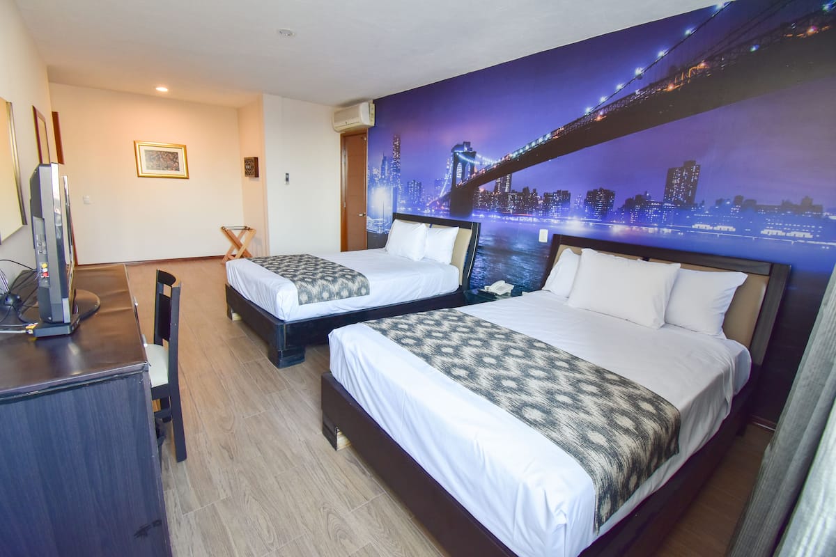 A hotel room with two beds, a desk with a computer, and a cityscape mural on the wall.