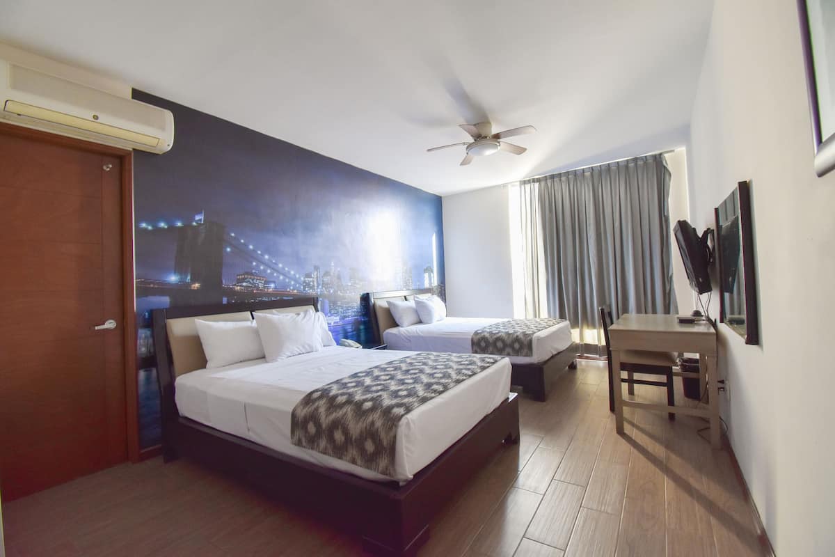 A hotel room with two beds, a desk, a ceiling fan, and a cityscape mural on the wall.