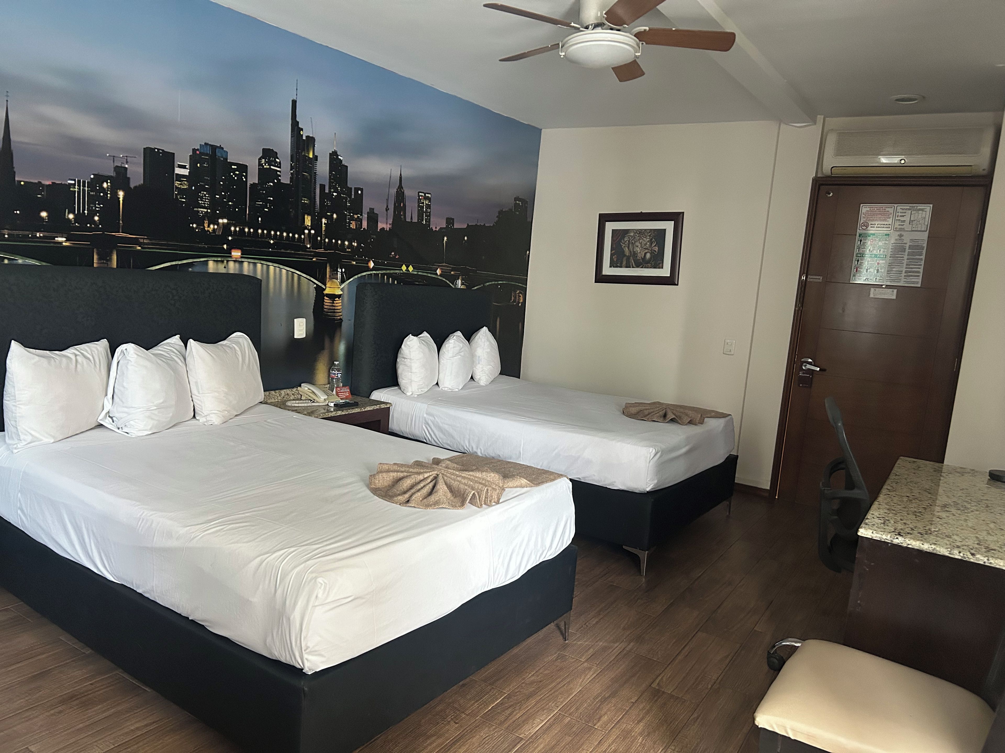 A hotel room with two beds, a cityscape mural, a ceiling fan, and a door with a notice.