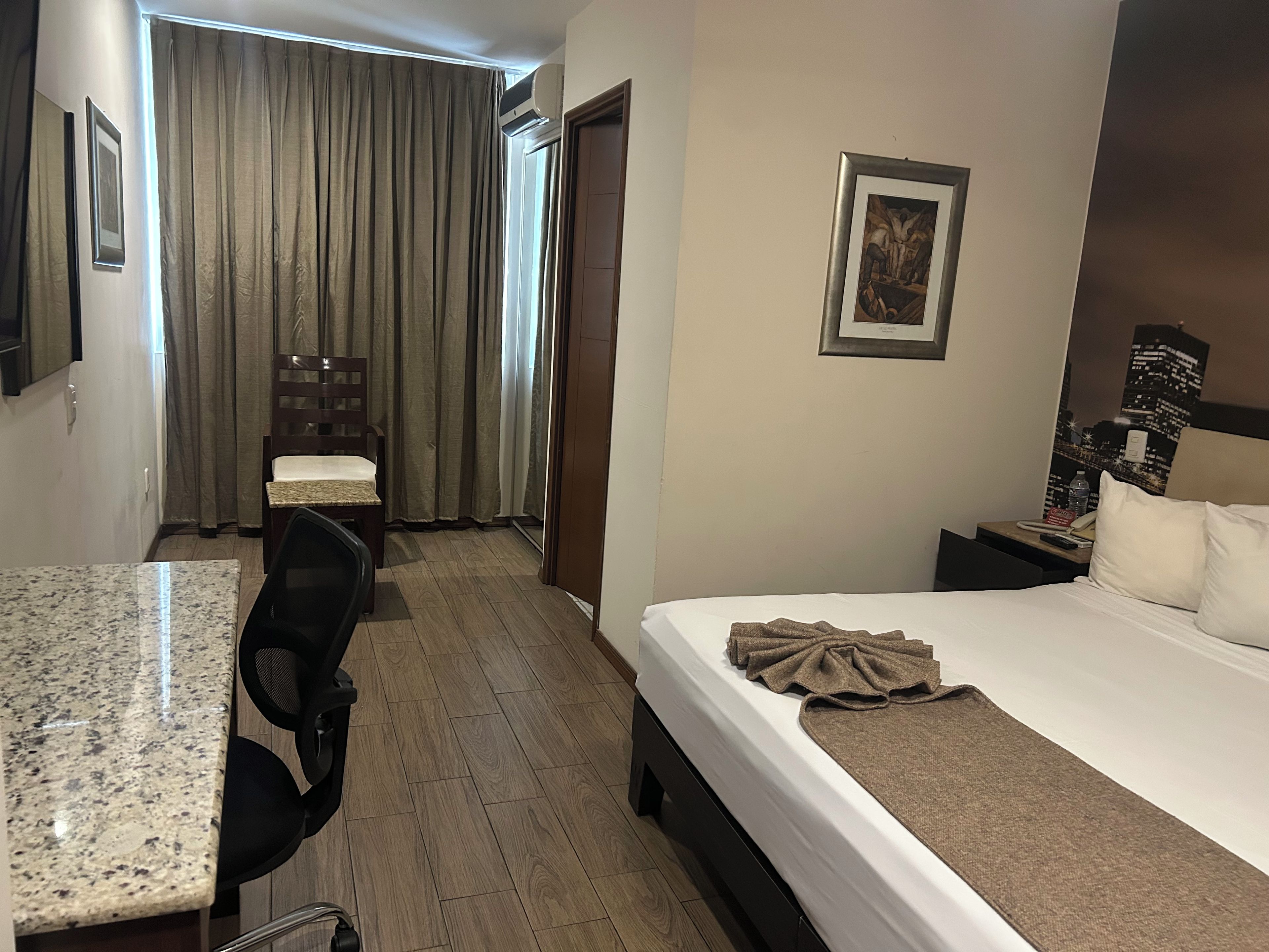 A hotel room with a bed, a desk, a chair, a window with curtains, and a painting on the wall.