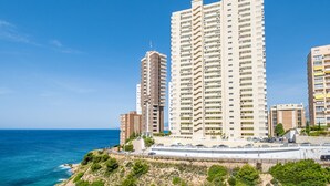 Een hoog appartementencomplex aan de kust met meerdere balkons, een parkeerplaats en uitzicht op zee.