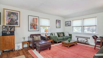 A living room with a brown leather sofa, a green sofa, a coffee table, and a red rug. There are framed pictures on the wall and a radio on a wooden cabinet.