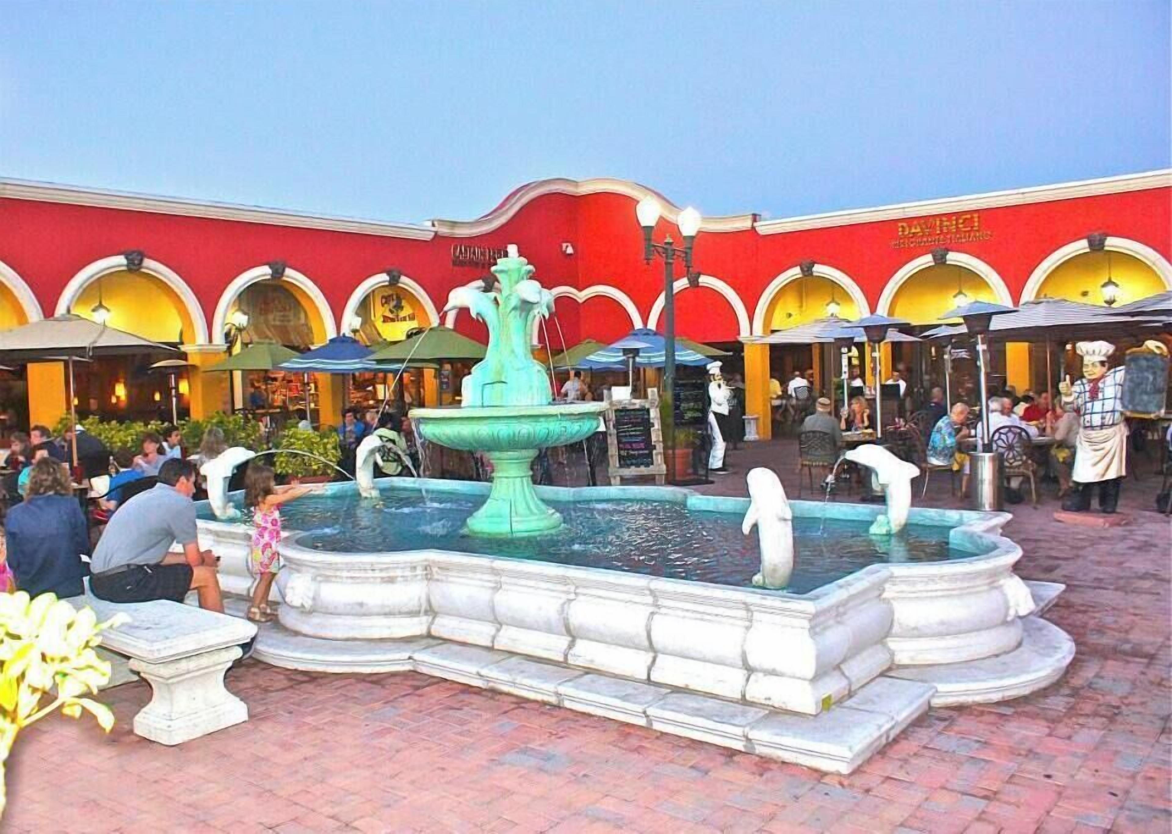 A public square with a central fountain, surrounded by outdoor seating areas and a building with multiple arches and a red facade.