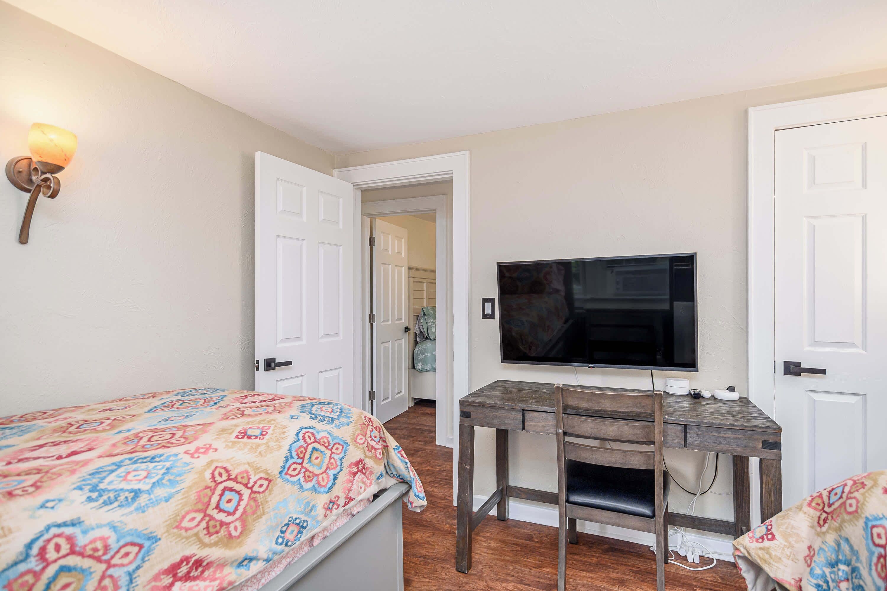 A bedroom with a bed, a desk with a chair, a television, and a door leading to another room.