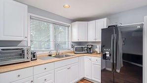 A modern kitchen with white cabinets, a stainless steel refrigerator, a microwave, a toaster, and a window with blinds. - Flex Comfort Apts of Greenwich #1 (Greenwich)