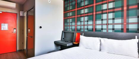 A hotel room with a bed, a desk, a red door, and a large window with a plaid pattern.
