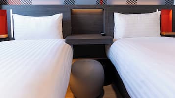 A hotel room with two beds, a bedside table with a remote control, and a round ottoman.