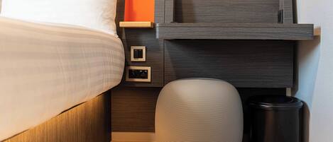 Un lit bien fait, avec des oreillers, une table de chevet sur laquelle se trouve une petite enceinte, une poubelle et une étagère fixée au mur, sur laquelle repose un objet d’un orange vif.