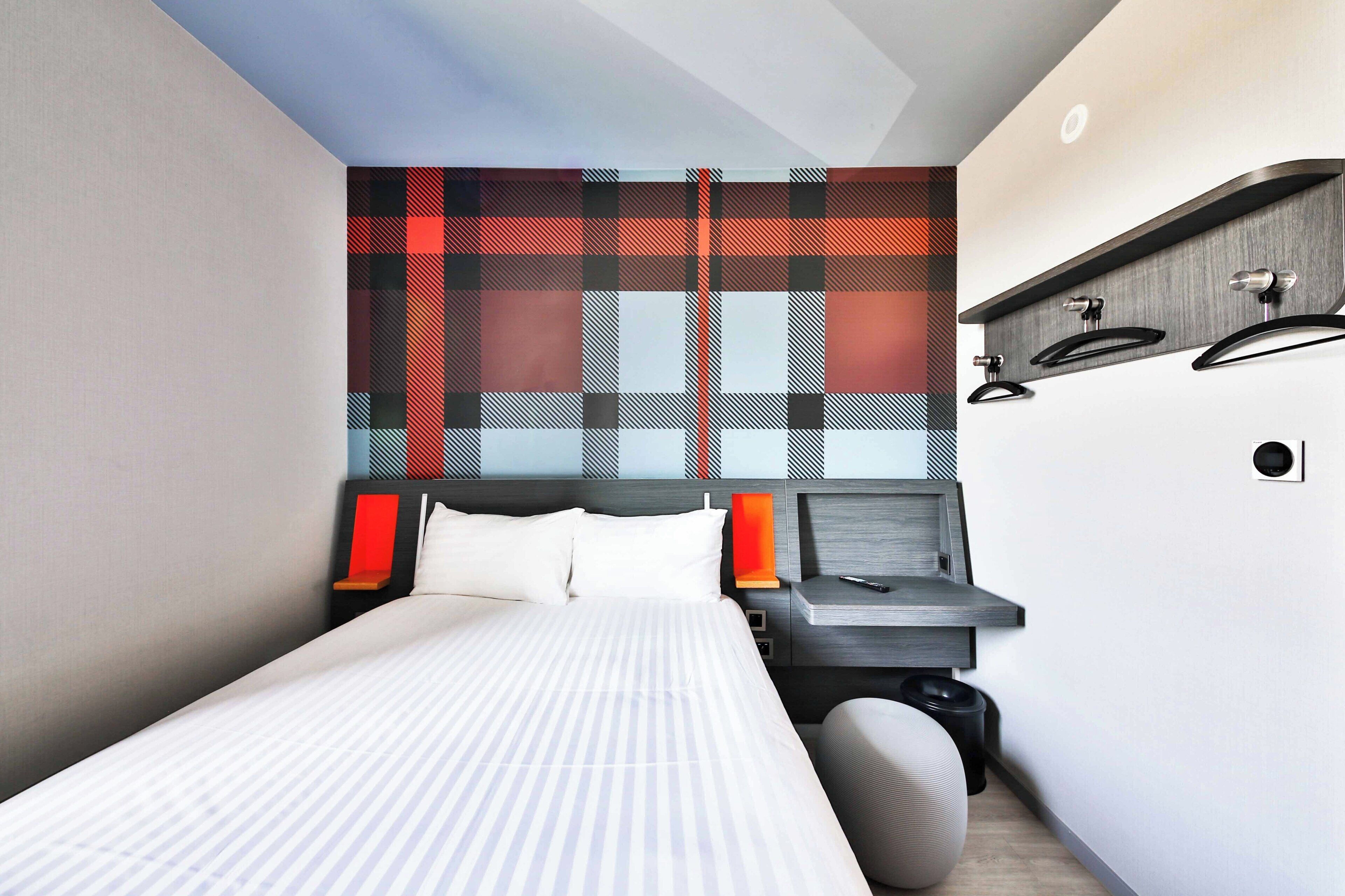 A modern hotel room with a bed, bedside table, and a wall-mounted shelf with a red lamp.
