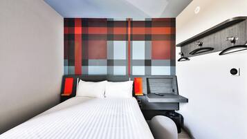 Une chambre d’hôtel moderne, équipée d’un lit, d’une table de chevet et d’une étagère fixée au mur, sur laquelle se trouve une lampe rouge.