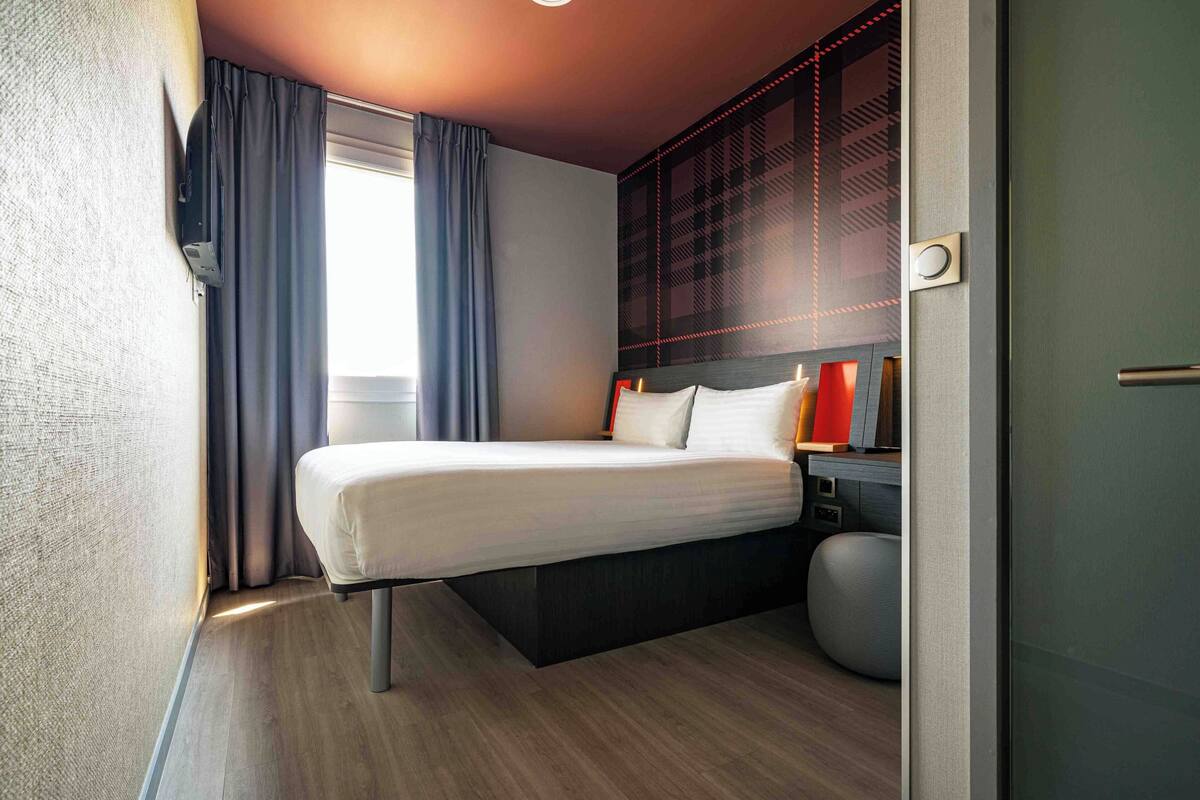 Ein modernes Hotelzimmer mit Bett, Nachttisch und großem Fenster mit Vorhängen.