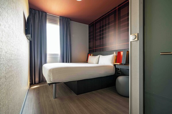 A modern hotel room with a bed, bedside table, and a large window with curtains. - easyHotel Paris Charles de Gaulle (Tremblay-en-France)