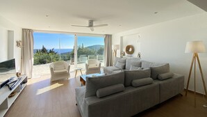 A modern living room with a grey sofa, a TV, and a floor lamp. There is a balcony with a view of the sea and hills. - Holiday rental villa with swimming pool in Begur, Casa de Campo (Begur)