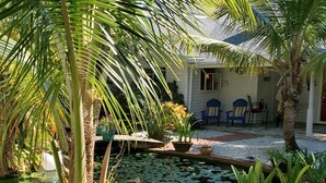 Un jardin paisible avec un étang, des palmiers et une maison dotée d’un espace extérieur aménagé pour s’asseoir.