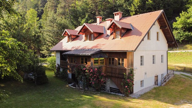 A traditional wooden house with a sloped roof, a balcony, and a small porch area with flowering plants.