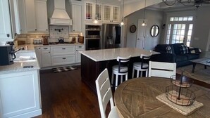 A modern kitchen with white cabinetry, a central island, and a dining area with a round wooden table. - Luxury by the Sea (Southold)