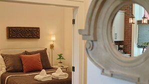 Una cama bien hecha con una colcha marrón y almohadas, una mesita de noche con una lámpara y una planta, y un espejo que refleja una pared de ladrillo y una ventana con vistas.