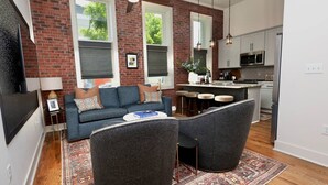 Una sala de estar moderna con un sofá, dos sillones, una mesa de centro y una zona de cocina con un microondas y taburetes de bar.