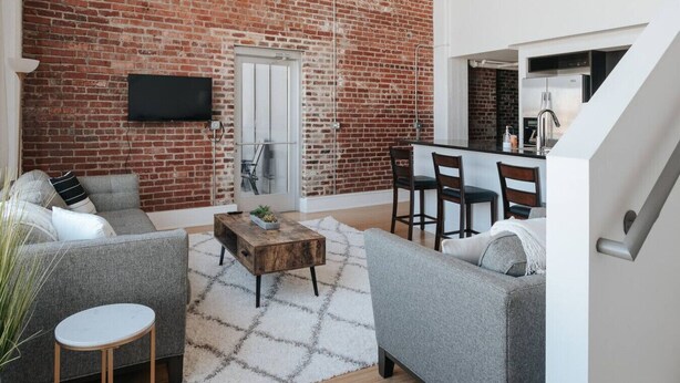 A living room with a brick wall, a flat-screen TV mounted above a door, a grey sofa, a wooden coffee table, and a small round table. - Glass Loft - Walk to Old City & DT Knox (Knoxville)