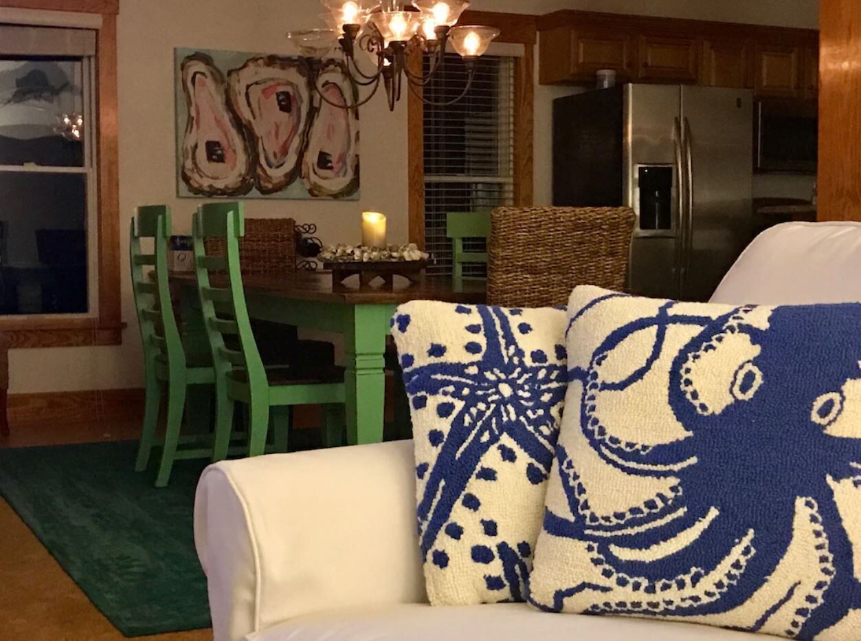 Un salon avec un canapé blanc, des fauteuils verts, un lustre et, au fond, une cuisine.