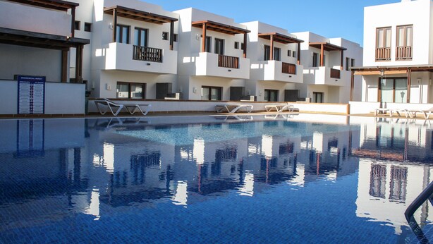 A swimming pool with clear blue water, surrounded by modern white buildings with balconies and wooden railings. - AIOLOS 20 by Buenavilla|Great apartment in the marina of Puerto Calero (Las Palmas)