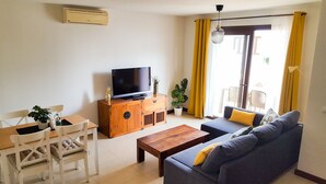 A living room with a sofa, a wooden coffee table, a TV on a wooden cabinet, a dining table with chairs, and a potted plant. - AIOLOS 20 by Buenavilla|Great apartment in the marina of Puerto Calero (Las Palmas)