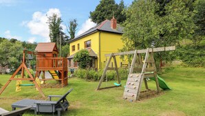 A playground with swings and slides, a yellow house, and a grassy area. - The Woodlands Lower Level (Narberth)