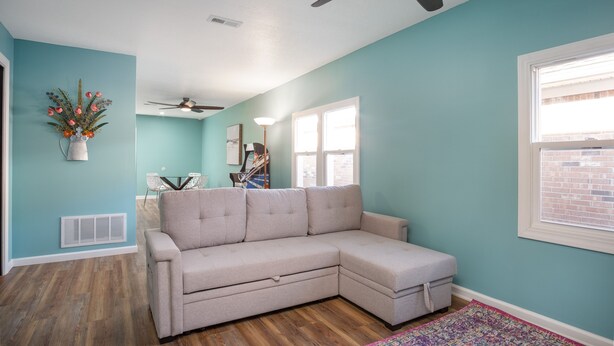 A room with a beige sectional sofa, a ceiling fan, a window with blinds, and a decorative plant.
