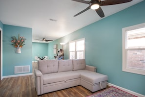Una habitación con un sofá seccional color beige, un ventilador de techo, una ventana con persianas y una planta decorativa.