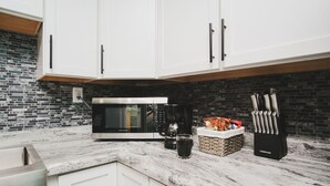 A kitchen with white cabinets, a black microwave, a coffee maker, a glass, and a knife block.