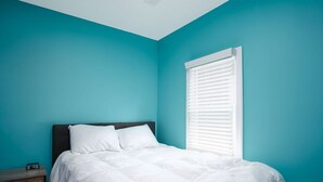 A bedroom with teal walls, a white bed, and a window with blinds.