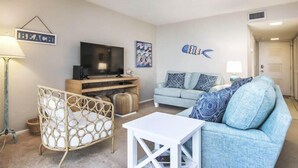 Ein Wohnzimmer mit Fernseher, einem blauen Sofa, einem weißen Tisch und einem Zierstuhl.