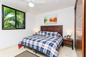 A bedroom with a bed, a ceiling fan, a nightstand with a lamp, a painting on the wall, and a window with a view of greenery.