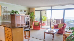 Un vaste espace de vie comprenant une cuisine, un fauteuil rouge, une table basse et une vue sur l’océan grâce à de grandes portes-fenêtres coulissantes.