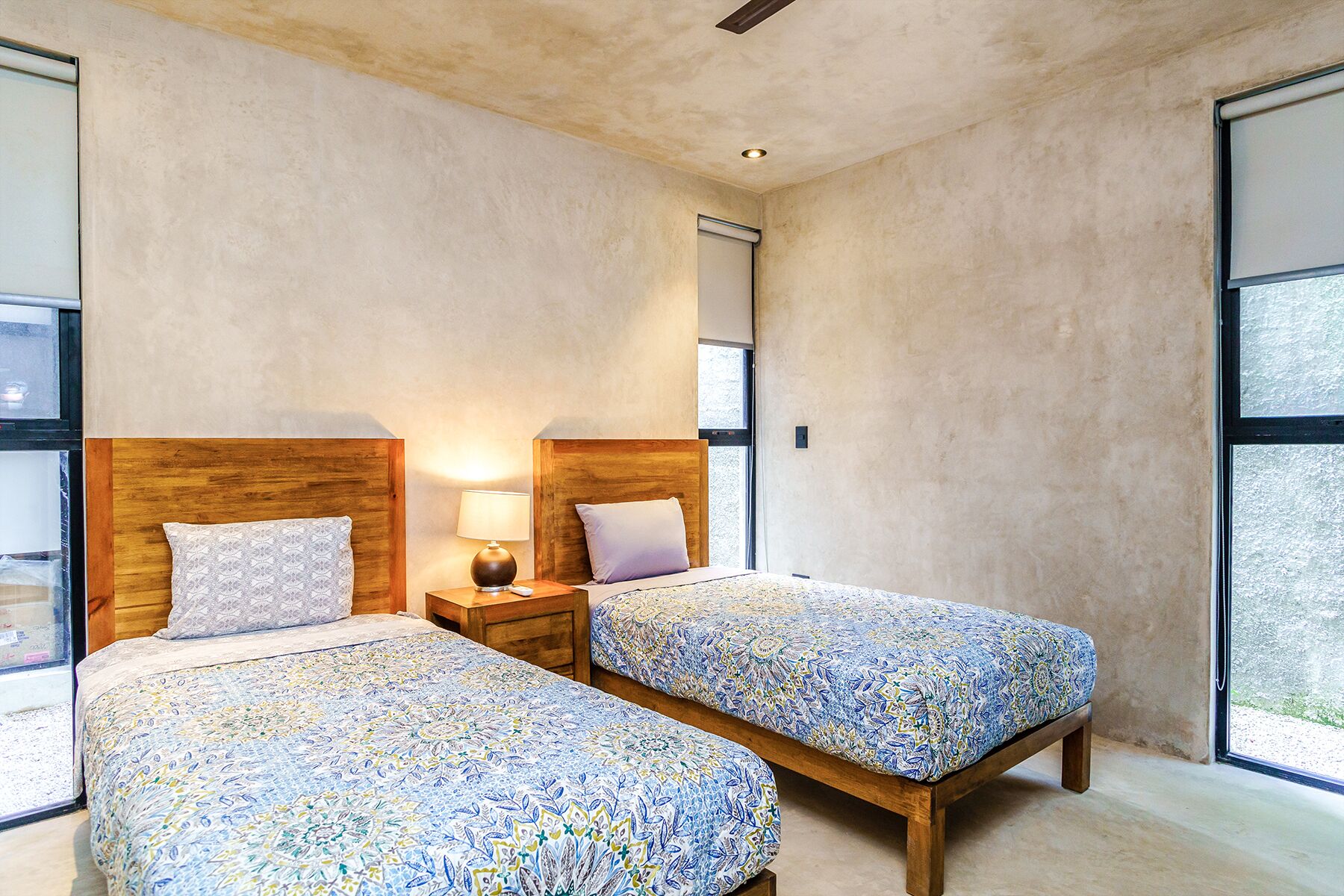 A bedroom with two beds, wooden headboards, bedside tables with lamps, and large windows.