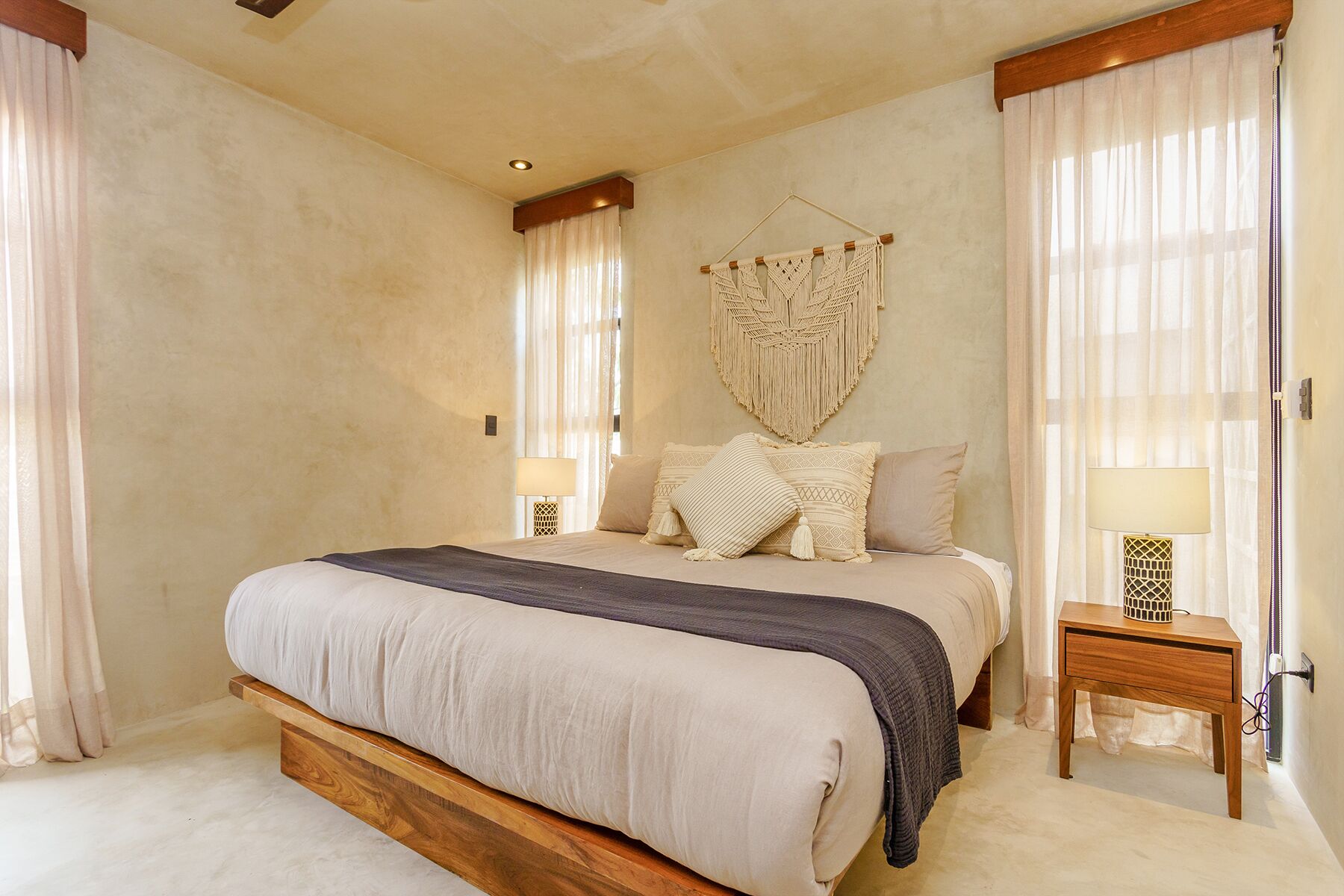 A bedroom with a large bed, two bedside tables with lamps, a macrame wall hanging, and sheer curtains.