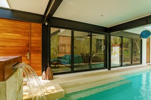 A modern indoor swimming pool with clear glass walls, a wooden deck, and a seating area with a green sofa and table. - Magazine-worthy Villa Ponderosa with Private Pool & Gated Area (Tulum)