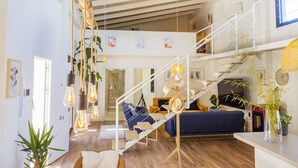Un amplio salón con una escalera de madera, un sofá azul y lámparas colgantes.