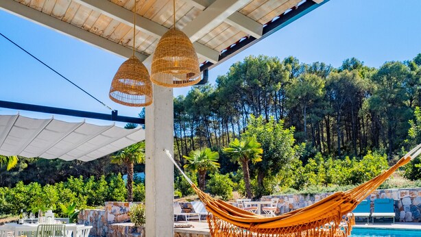 Un espacio exterior con asientos de bambú y una hamaca, con vista a una piscina y una zona boscosa.