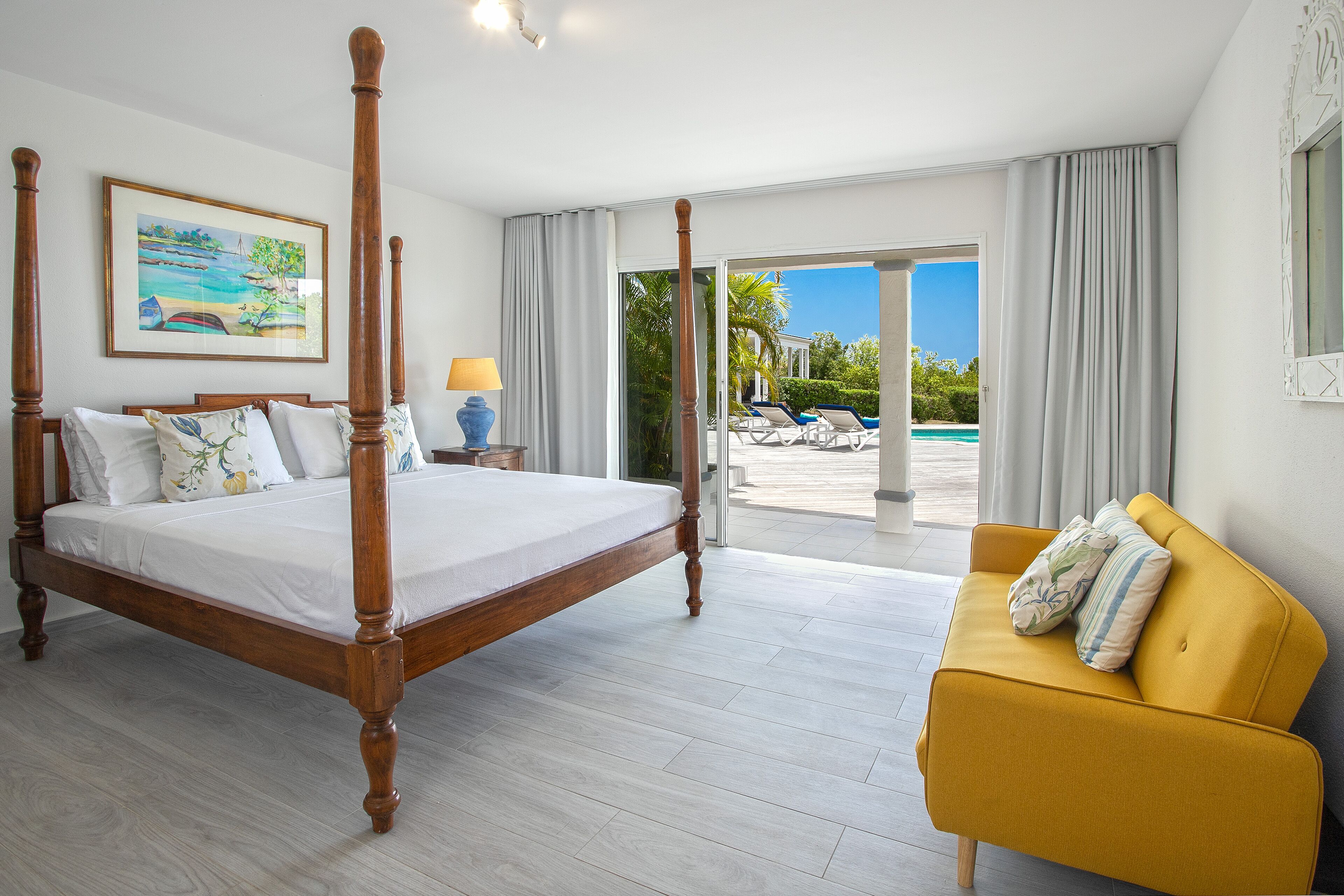 Ein Schlafzimmer mit Himmelbett, gelbem Sofa und einem großen Fenster mit Blick auf einen Pool und Palmen.