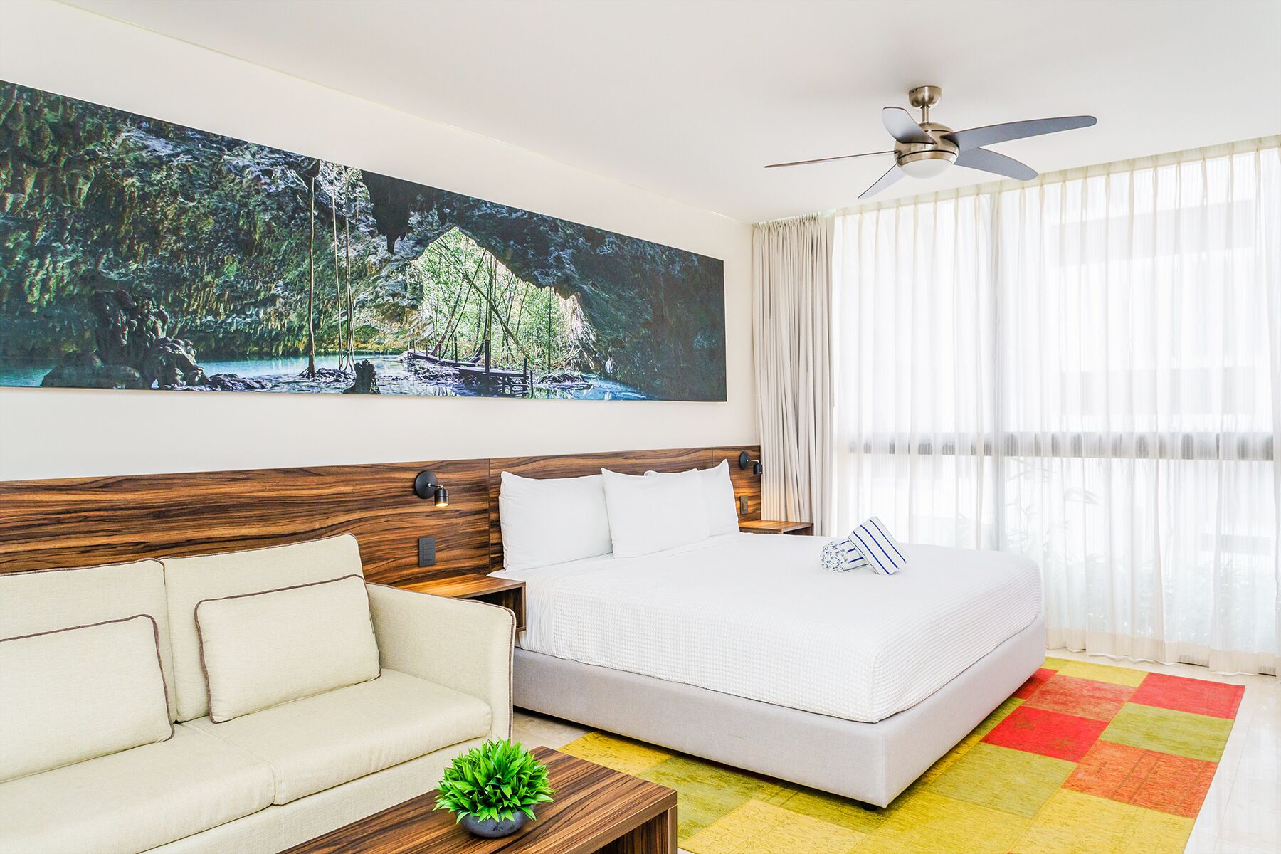 A hotel room with a bed, a sofa, a ceiling fan, and a large painting of a forest scene.